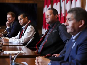 Four men are seen sitting at a table with microphones in front of canadian flags.