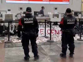 Police officers stand in a bank lobby as protesters sit near teller desks with a banner and papers strewn on the floor