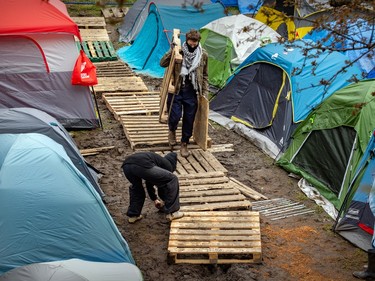 Two men build a walkway out of wooden pallets between two rows of tents on muddy terrain