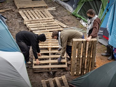 Three people lay down wood pallets to create a walkway on muddy terrain between two rows of tents