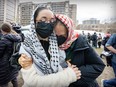 Two women wearing Palestinian scarves and masks hug outside, with other people and media visible in the background