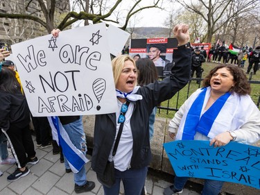 Two women hold homemade signs reading 'We are NOT afraid!' and 'Montreal stands with Israel' while yelling in front of a fence. Police and Palestinian flags are visible in the distance.