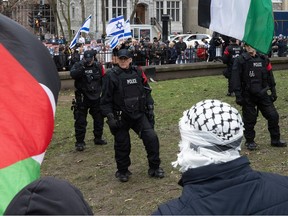 Police officers wearing tactical gear and red armbands stand between people carrying Palestinian flags on the lawn and people carrying Israeli flags beyond a fence on the sidewalk