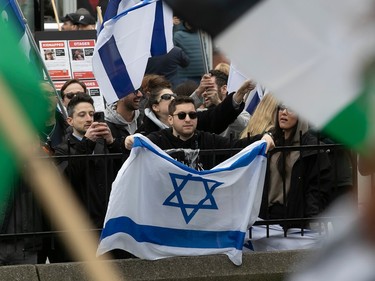 A man holds an Israeli flag over a fence surrounded by other people carrying Israeli flags and hostage posters, while Palestinian flags are visible blurry in the foreground