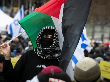 A person wearing a headscarf and face covering holds a Palestinian flag behind their head, with Israeli flags in the background