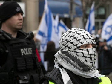 A person wearing a kaffiyeh as a headscarf and face covering stands in front of a police officer, both facing the same direction, with Israeli flags in the background