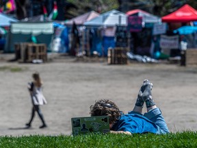 A young person lies down on the grass with a laptop, with an encampment visible in the background