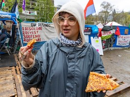 Ari Nahman, wearing a rain jacket and hoodie, holds two slices of pizza while speaking outside an encampment