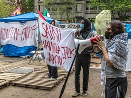 Two masked protesters hold up a banner while a third speaks into a megaphone at an encampment with wooden pallets on the ground.