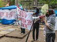 Two masked protesters hold up a banner while a third speaks into a megaphone at an encampment with wooden pallets on the ground.
