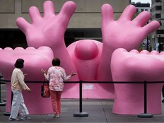 Two women stand in front of a giant pink statue of a cartoonish person with their hands and feet in the air.
