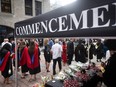 People, some in robes, are visible behind a pop-up tent with the word 'Commencement' on it and flowers and other items on a table