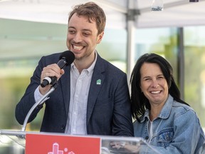 A smiling man grabs a microphone on a podium outdoors while Valérie Plante grabs his arm