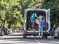 A man lowers a chair out of a moving van while another man sorts through boxes in the van in the middle of a road.