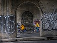 Two people wearing yellow tops ride on bike-share bikes through a tunnel. They're frame by a cement arch with graffiti on either side.
