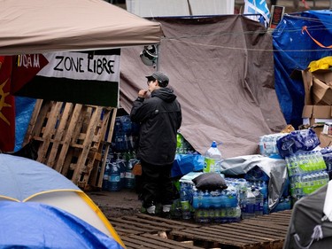 A person is seen brushing their teeth inside an encampment, while stacks of new water bottles lie on the floor nearby