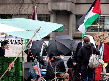 People gather at the entrance to an encampment, with several open umbrellas and Palestinian flags
