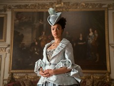 A woman in Regency costume and hat stands in front of an oil painting.
