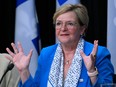 Guylaine Leclerc raises her hands while speaking behind a microphone with Quebec flags in the background