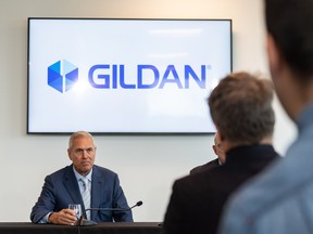 A man in a suit sits at a table with the logo of Gildan on a screen behind him