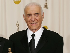 A man in a black suit and tie smiles in front of a backdrop with Oscar statuettes