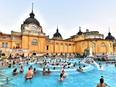 Dozens of people are seen enjoying the hot springs-fed baths at the Szechenyi Baths in Budapest in front of a majestic-looking building with a yellow facade.