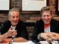 Bobby Slayton and Andy Nulman sit at a restaurant table next to a brick wall with framed pictures