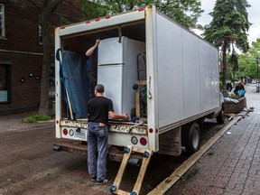 Two men unload a fridge from a moving van on a leafy street. One is inside the van, the other is outside the van and about to lift the fridge from the bottom.