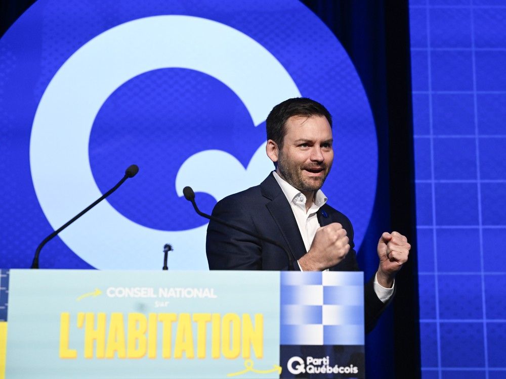 A man smiles and gestures during a press conference. The PQ logo is behind him.