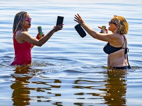 Two women in a lake, facing each other, holding a drink can in one hand and an electronic device in the other