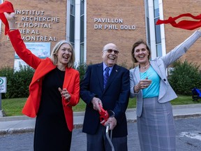 A man with a cane, centre, has just cut a ribbon. Two women beside him hold the ribbon and smile. Behind is a sign on a building that says Pavillon Dr Phil Gold