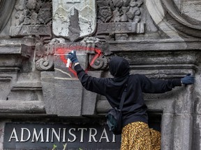 A person wearing a black hoodie and gloves spraypaints a red triangle on a sculpture on a stone wall above the word 'Administration'