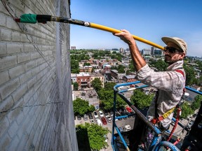 A man on a cherrypicker uses a pole to paint on a brick wall high up in the air