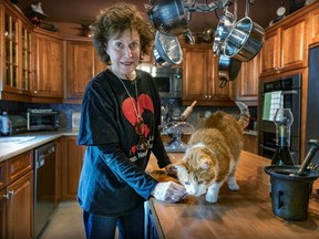 A woman stands next to an orange cat who is standing on a kitchen counter.