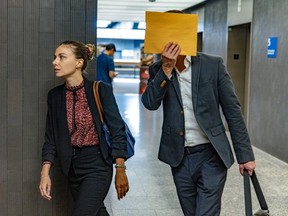 A woman and a man are seen in a courtroom hallway. The woman is facing the left frame as she turns out of the hallway, the man his hiding his face with a large envelope.
