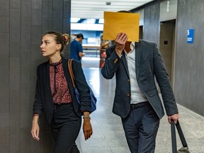 A woman in a courthouse hallway walks beside a man holding a manilla envelope over his face.