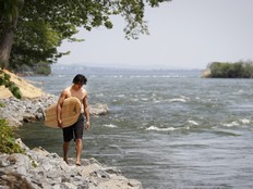 A man carries a surfboard along the shore of a river on the left side of the frame, there's a river with rapids to the right.
