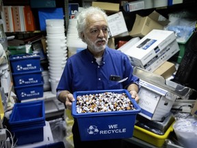 A man holds a recycling bin full of empty medicine vials. There are many boxes behind him.
