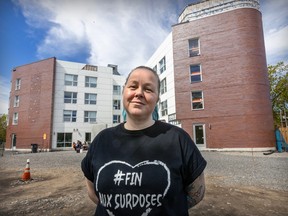 A woman smiles in front of a building and a gravel lot. She is wearing a t-shirt that reads Fin Aux Surdoses.