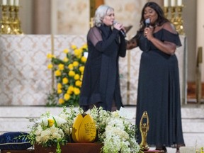 Two women sing behind a casket topped with flowers
