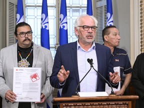 A man speaks behind a podium, flanked by two other men with Quebec flags in the background