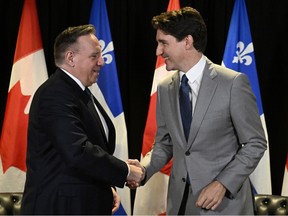 François Legault and Justin Trudeau shake hands in front of Quebec and Canadian flags, Trudeau smiling and Legault with more of a smirk