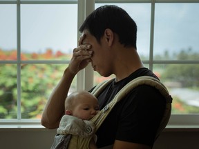 A depressed dad at home with his child covering his face and standing next to the window.