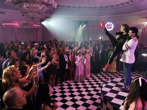 A crowd of well-dressed people watch as a man in a retro sweater holds up a woman dressed in black and shiny red boots doing a high kick in the air in a ballroom with a checkered floor