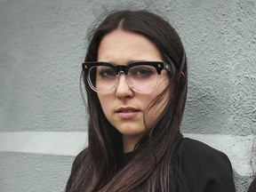 A woman with brown hair and dark glasses looks into the camera standing against a wall