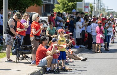 A large group of people on a sidewalk look at the roadway during a Canada Day parade.