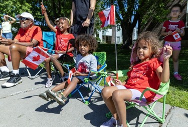 Children smile as they sit in chairs on a sidewalk and watch a Canada Day parade.