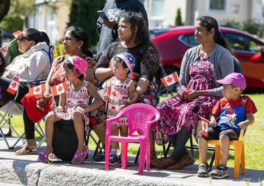 A group of women and children sit by the roadside while watching a Canada Day parade.