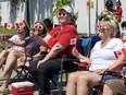 Four women wearing sunglasses shaped like Canadian flags smile and sit while waving flags as they watch a Canada Day parade.