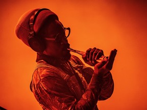 André 3000 plays flute while wearing headphones against a bright orange background.